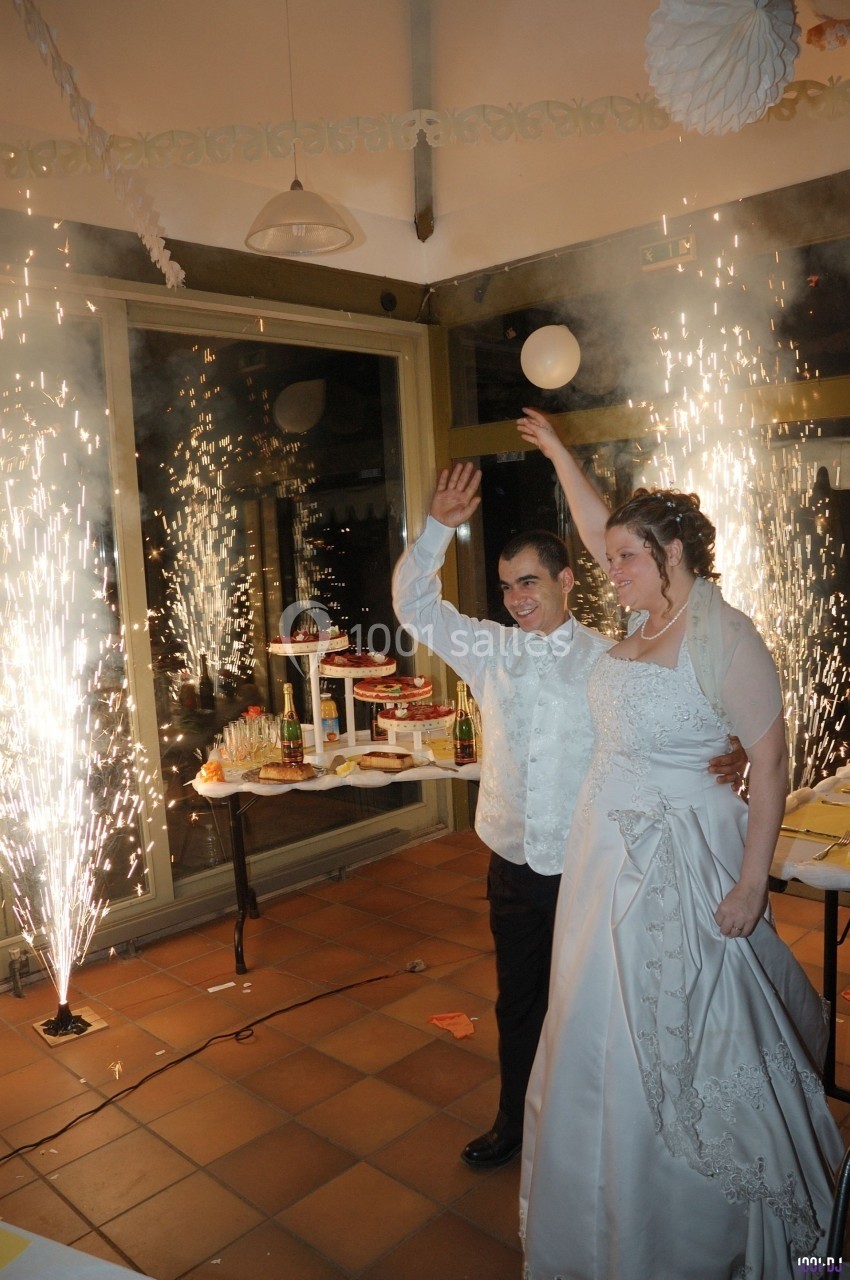 Un couple en tenue de mariage salue devant des étincelles lors d'une réception festive en intérieur.