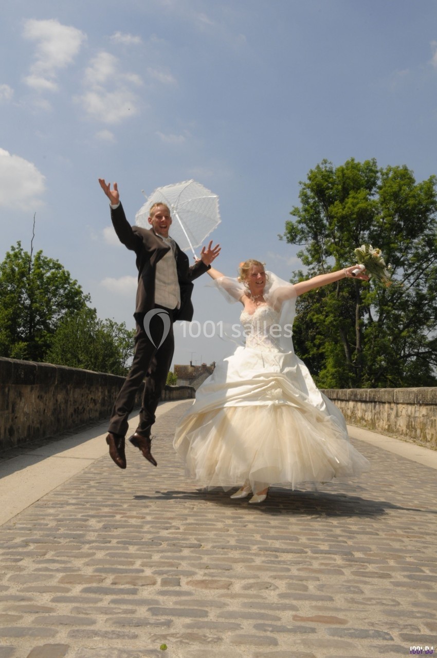 Un couple en tenue de mariage saute joyeusement sur un pont pavé par une journée ensoleillée.
