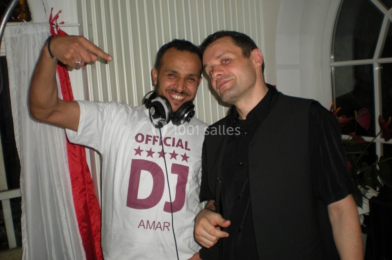 Deux hommes posent devant un décor intérieur, l'un portant un casque audio et un t-shirt ’Official DJ’.