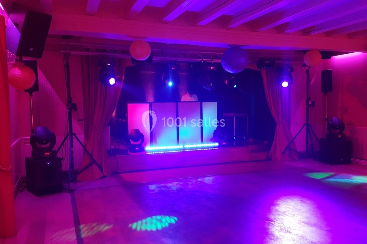 Salle de réception éclairée par des lumières colorées, avec une scène équipée pour un DJ et des enceintes.