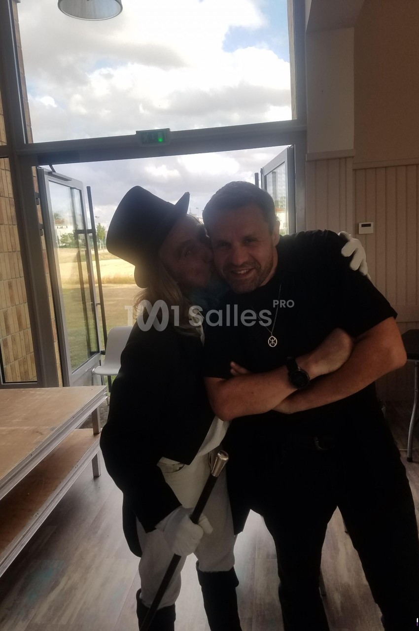 Un homme en costume élégant avec un chapeau embrasse un autre homme souriant dans un espace intérieur lumineux.