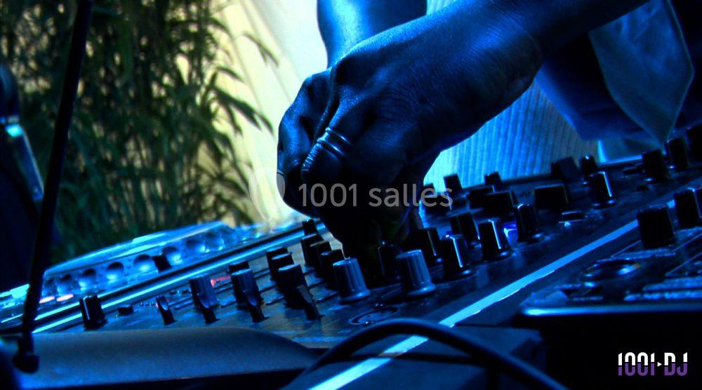 Main posée sur une table de mixage avec des boutons et des curseurs, éclairée par une lumière bleutée.