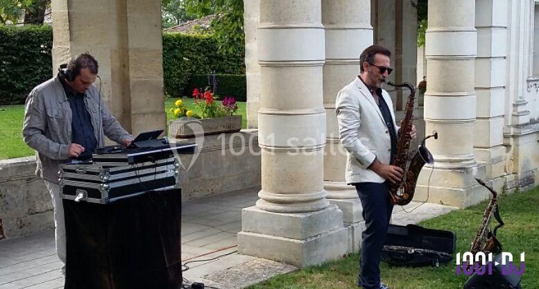 Un saxophoniste joue devant des colonnes en pierre, accompagné d'un DJ utilisant une table de mixage en extérieur.