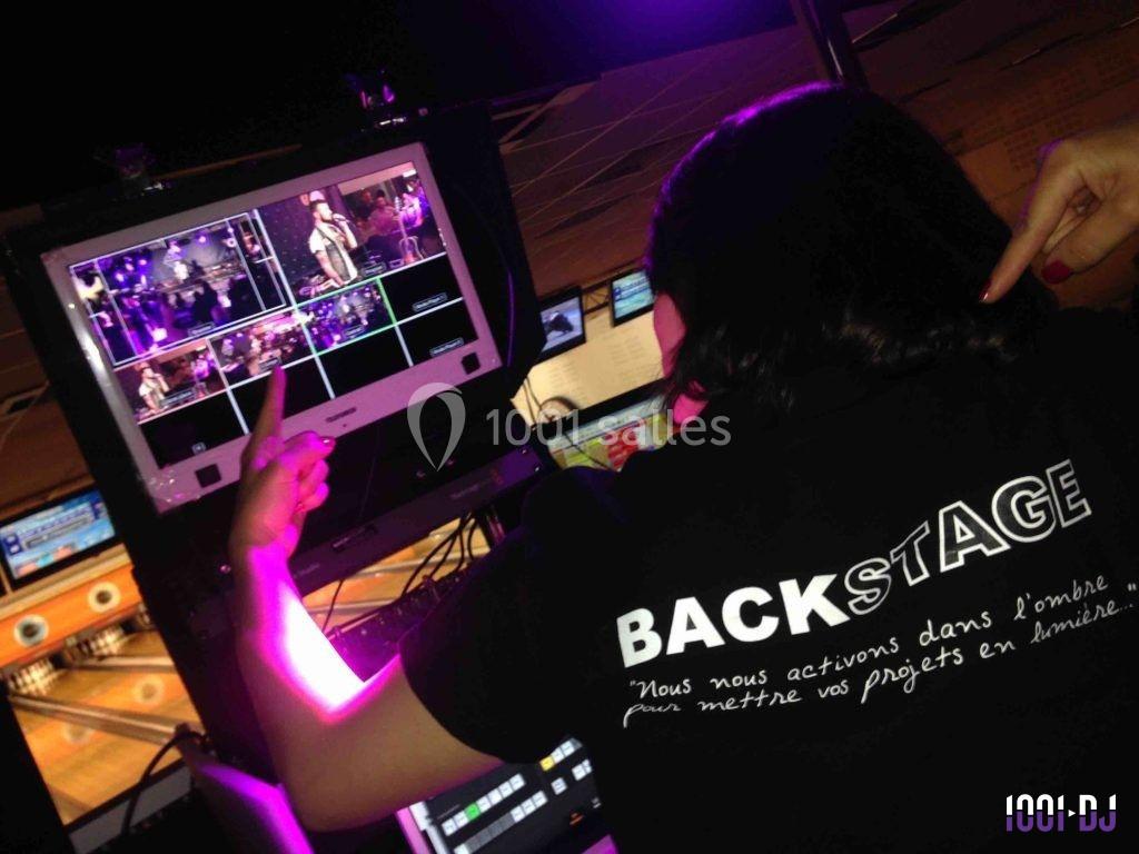 Une technicienne supervise des écrans de contrôle en coulisses, portant un t-shirt avec l'inscription ’Backstage’.