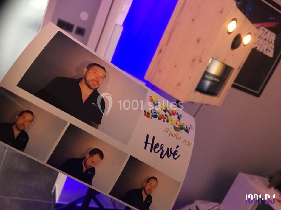 Une main tient une planche de photos imprimées d'un homme devant un photomaton en bois dans une pièce éclairée.