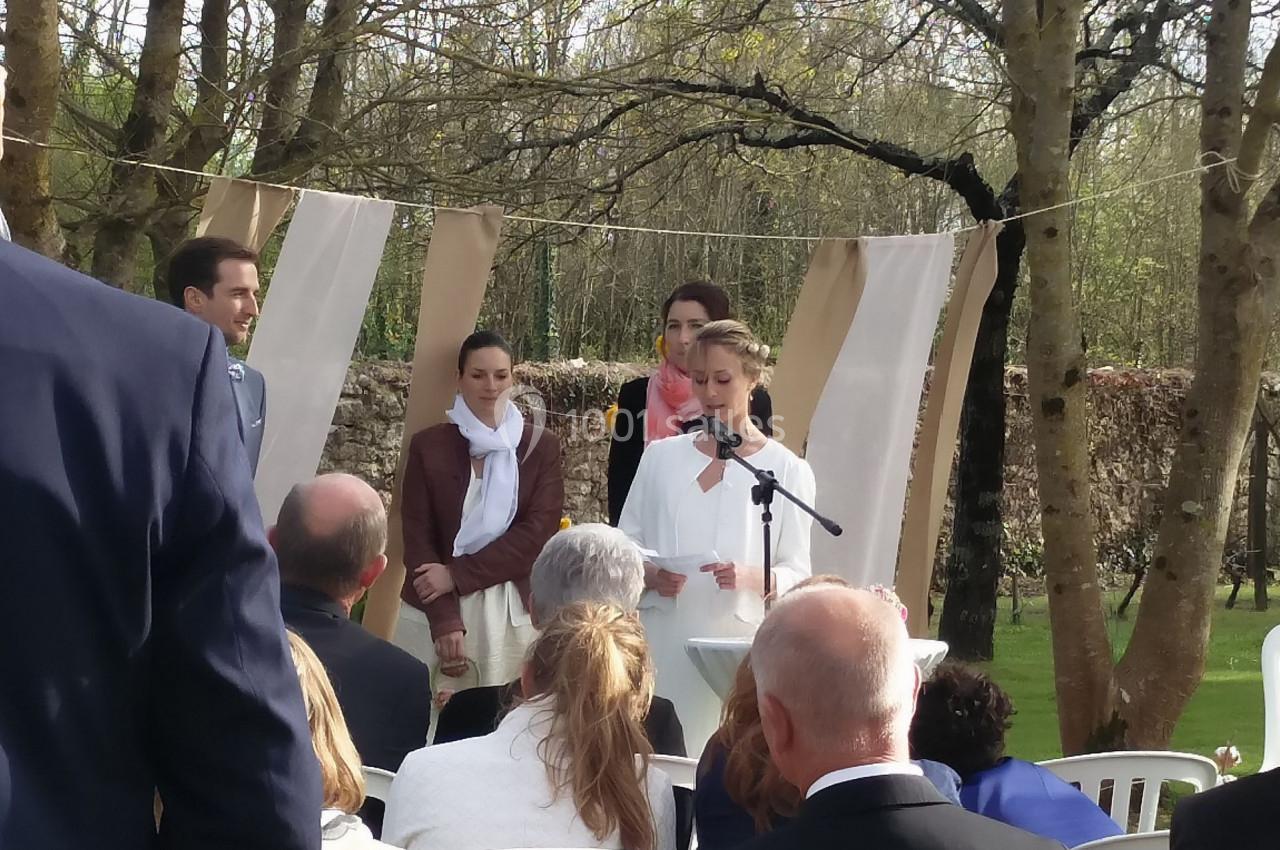 Une femme lit un discours au micro lors d'une cérémonie en plein air, entourée d'invités assis et de décorations.