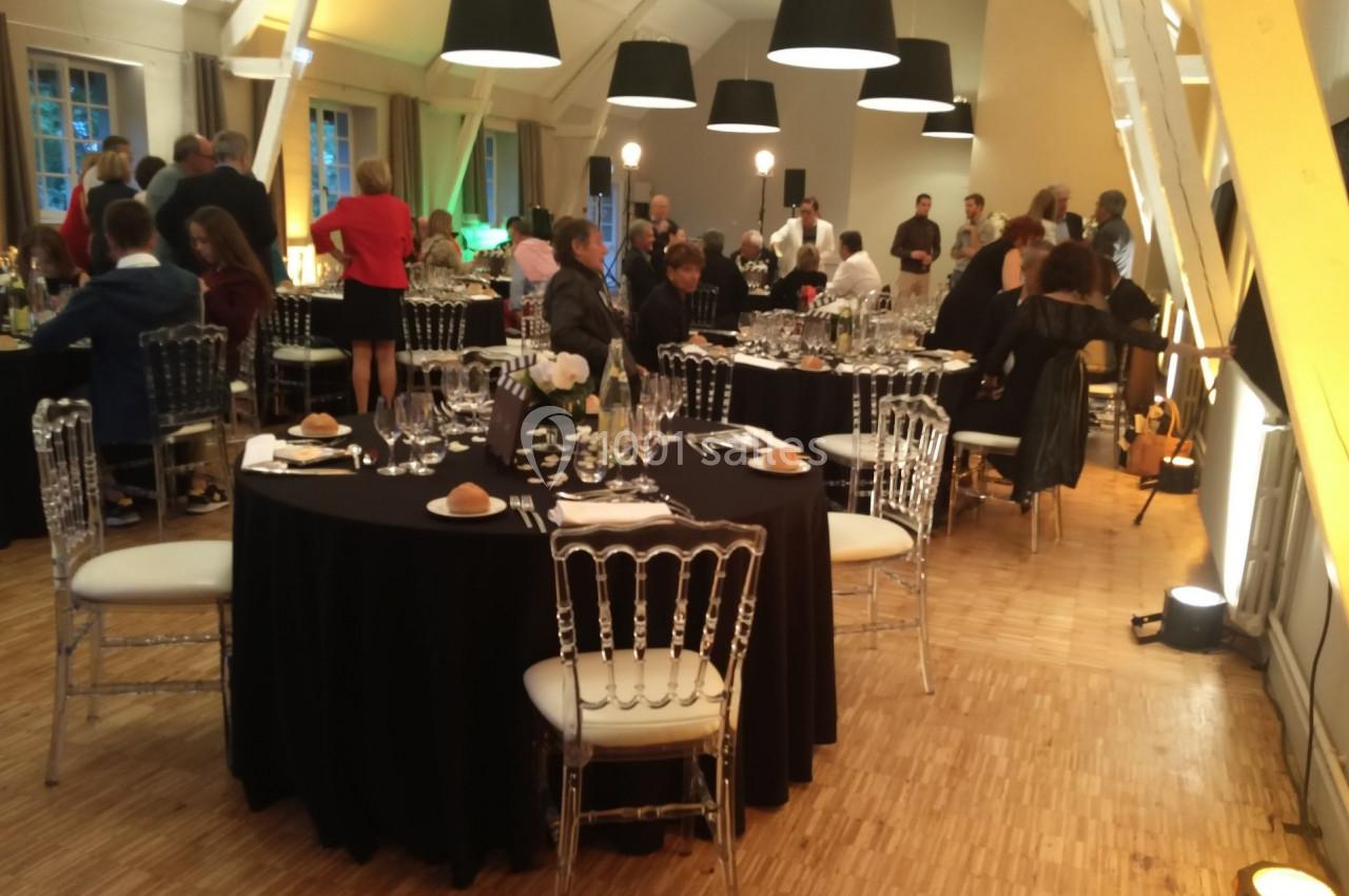 Salle de réception avec tables rondes dressées, invités debout et assis, éclairage chaleureux et ambiance conviviale.