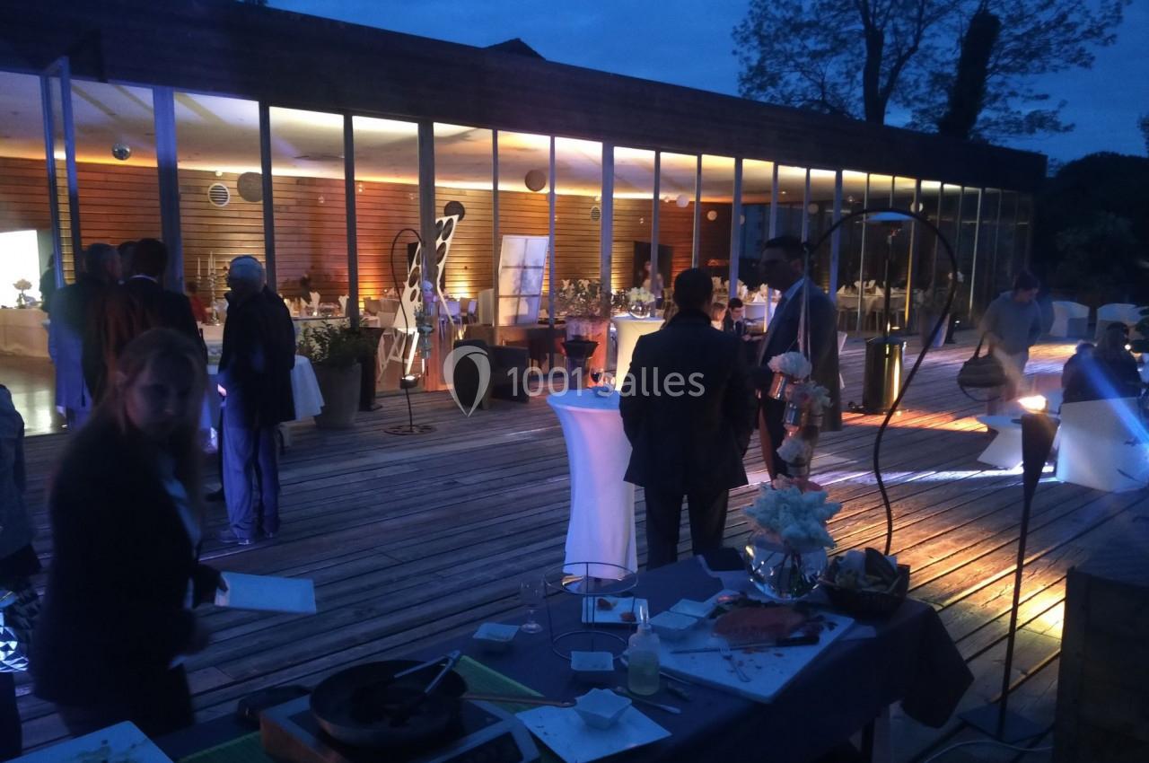 Personnes rassemblées sur une terrasse en soirée, devant une salle vitrée illuminée, ambiance conviviale.