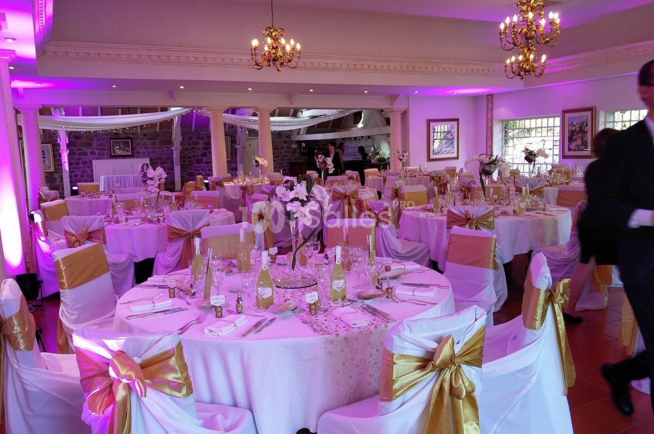 Salle de réception décorée avec des tables rondes, nappes blanches, chaises ornées de nœuds dorés et éclairage rose.