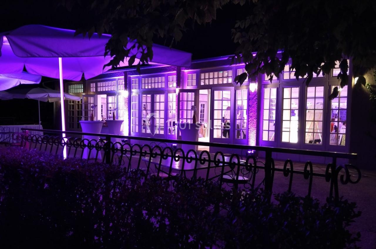 Façade d'un bâtiment éclairé en violet la nuit, avec des parasols et une clôture au premier plan.