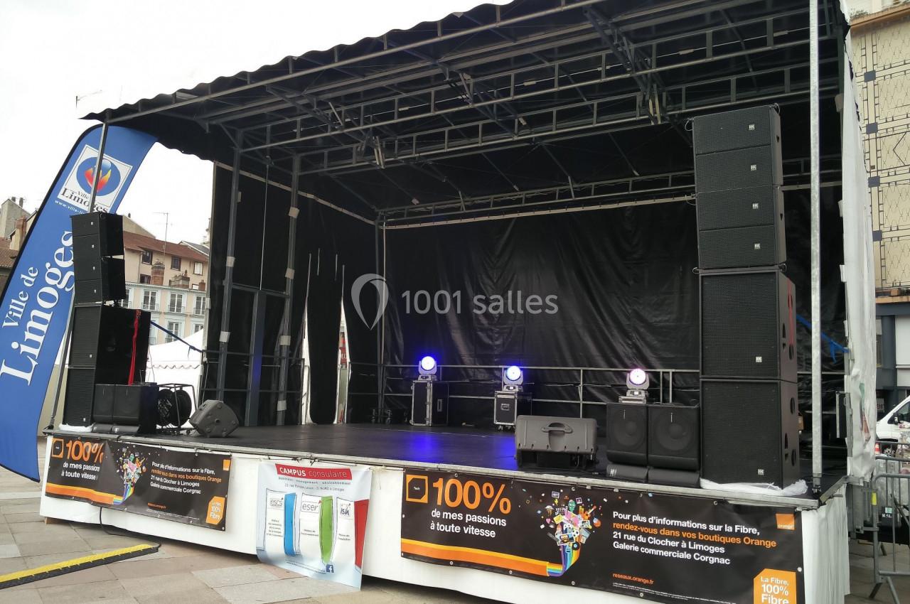 Scène extérieure vide avec équipements de sonorisation, située sur une place en ville, entourée de panneaux publicitaires.