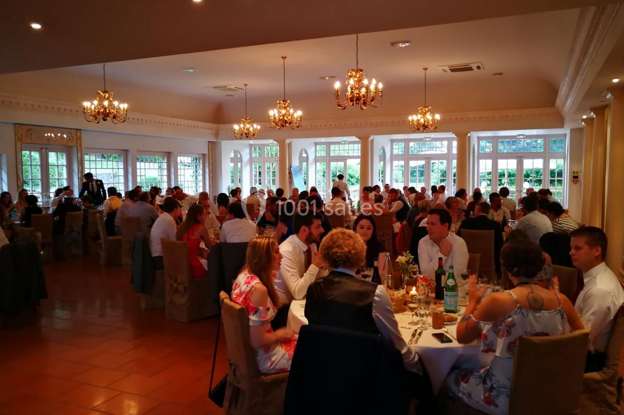 Salle de réception lumineuse avec des invités assis à des tables rondes, décorée de lustres et grandes fenêtres.