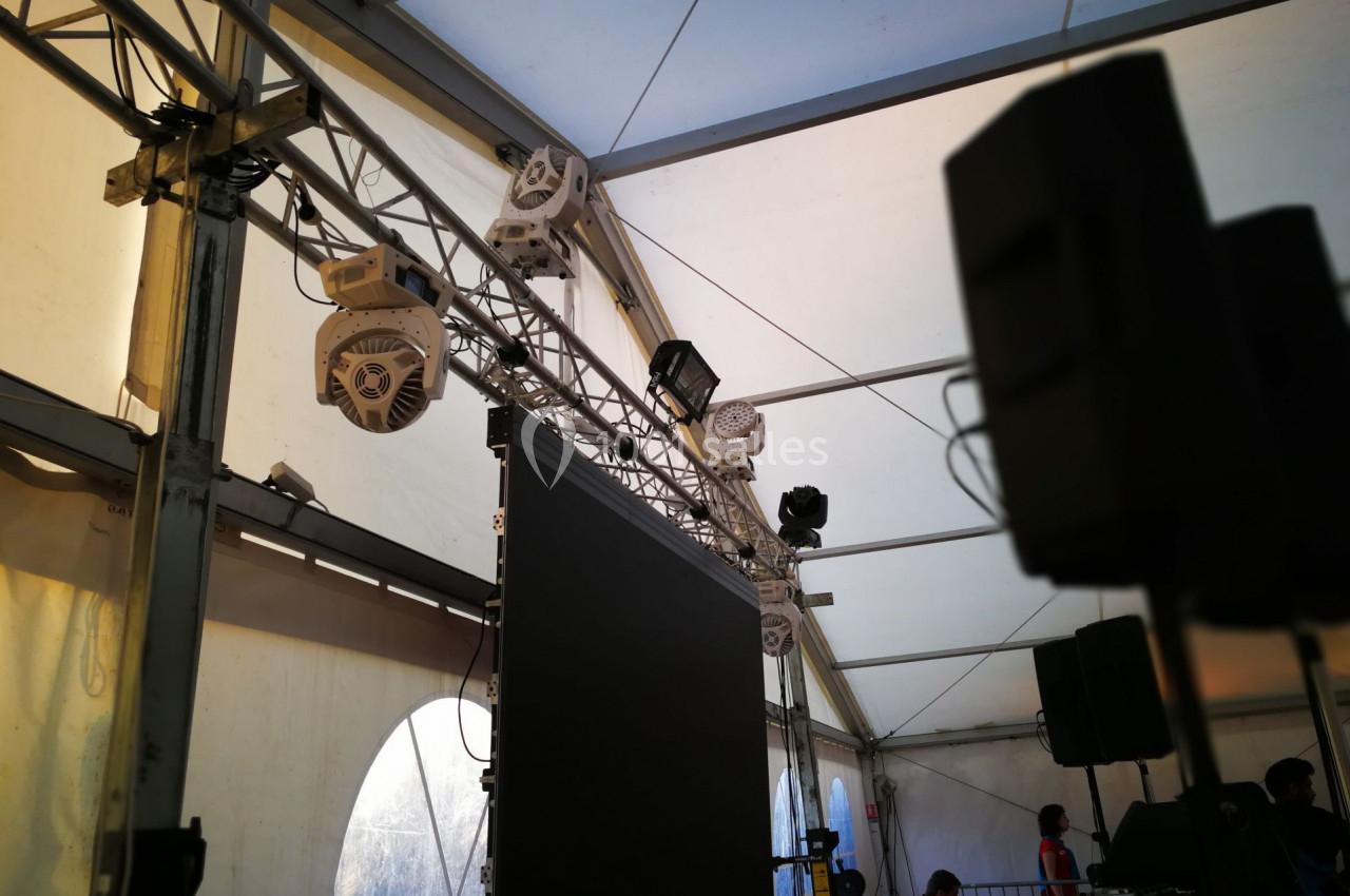 Structure métallique avec projecteurs, écran suspendu et haut-parleurs dans une tente événementielle éclairée.