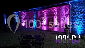 Façade en pierre éclairée par des lumières violettes et bleues, avec des tables en métal noir au premier plan.