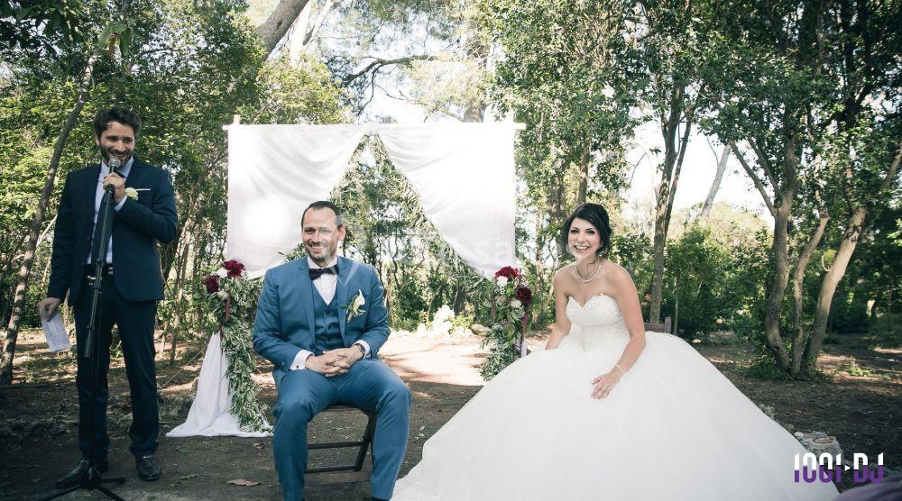 Un couple assis lors d'une cérémonie de mariage en extérieur, avec un officiant debout à côté d'une arche décorée.
