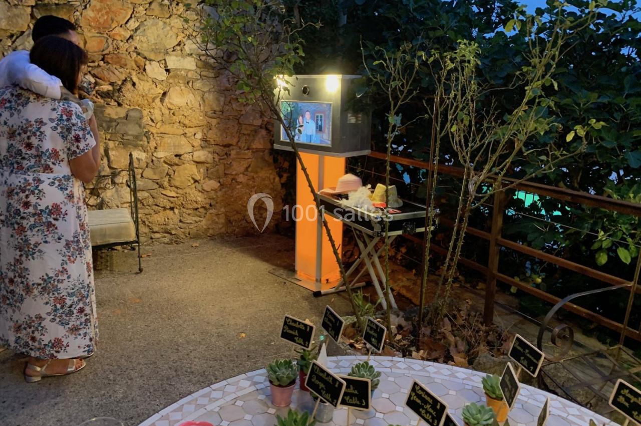 Un couple regarde un photobooth lumineux près d'un mur en pierre, avec une table décorée de plantes en premier plan.