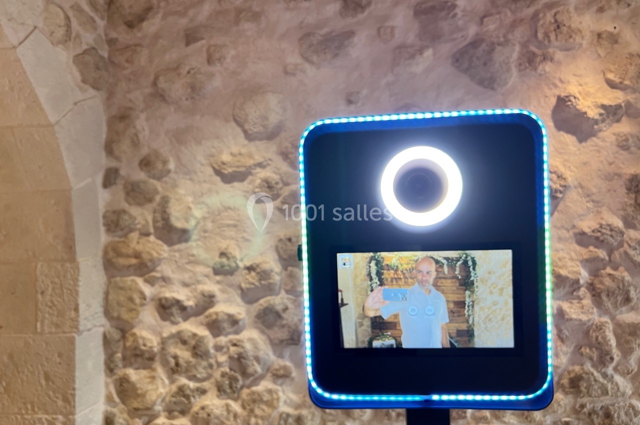 Borne photo avec écran affichant une personne posant, installée devant un mur en pierre dans un espace intérieur.