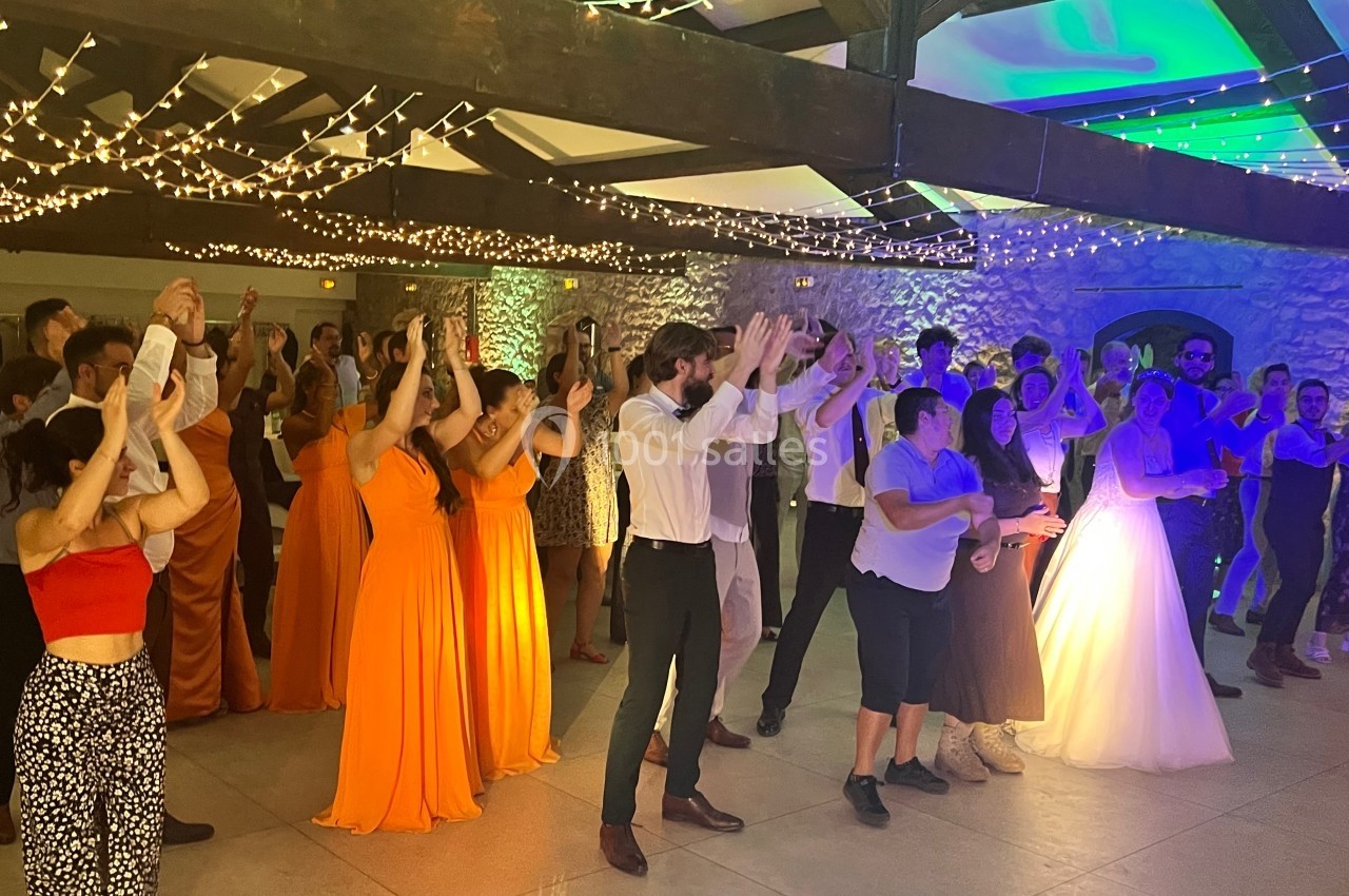 Groupe de personnes dansant sous des guirlandes lumineuses dans une salle avec murs en pierre, dont une mariée en robe…