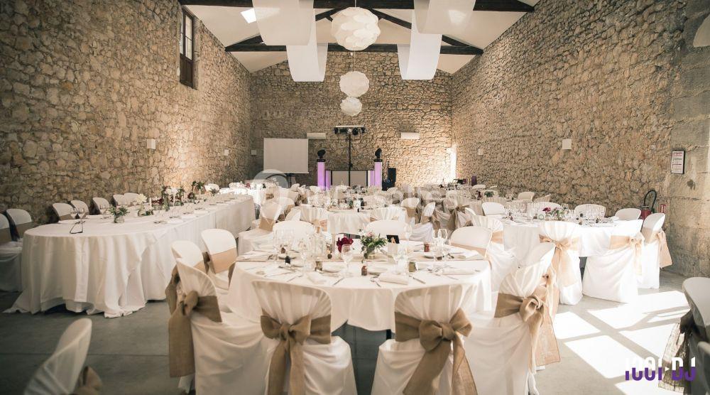Salle de réception en pierre avec tables rondes décorées de nappes blanches et chaises ornées de nœuds beiges.