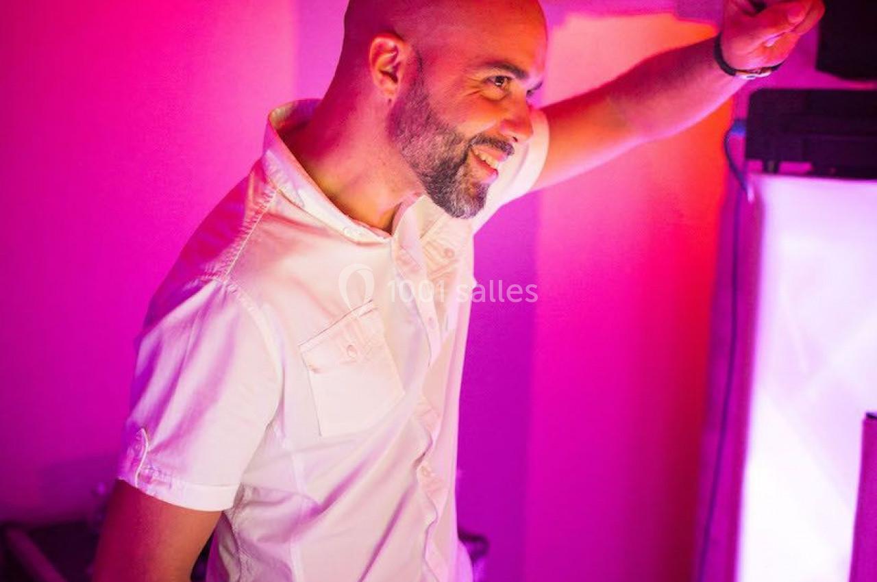 Un DJ souriant en chemise blanche mixe de la musique dans une ambiance lumineuse rose et violette.