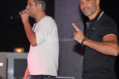 Deux hommes sur scène, l'un chante au micro tandis que l'autre pointe du doigt près d'un équipement audio.