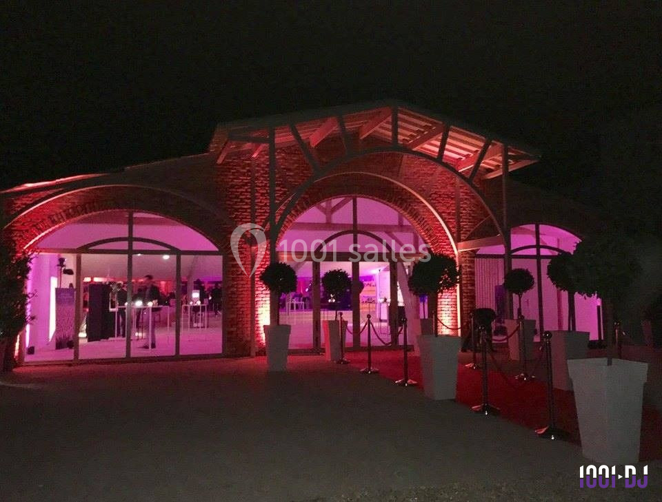 Façade d'une salle de réception éclairée en rose, avec tapis rouge et décorations végétales en soirée.