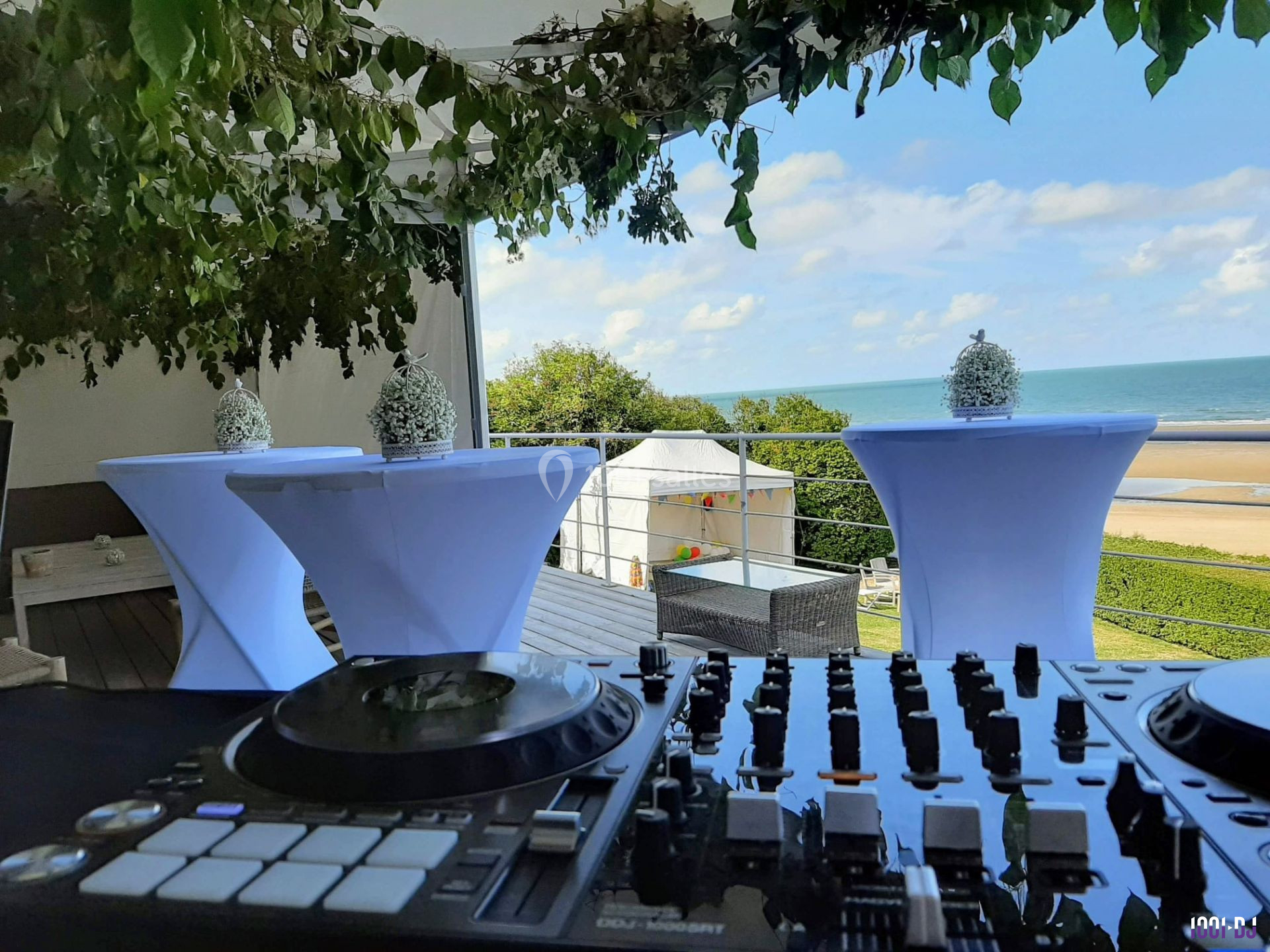 Vue d'une table de mixage en premier plan avec une terrasse décorée, donnant sur une plage et la mer en arrière-plan.