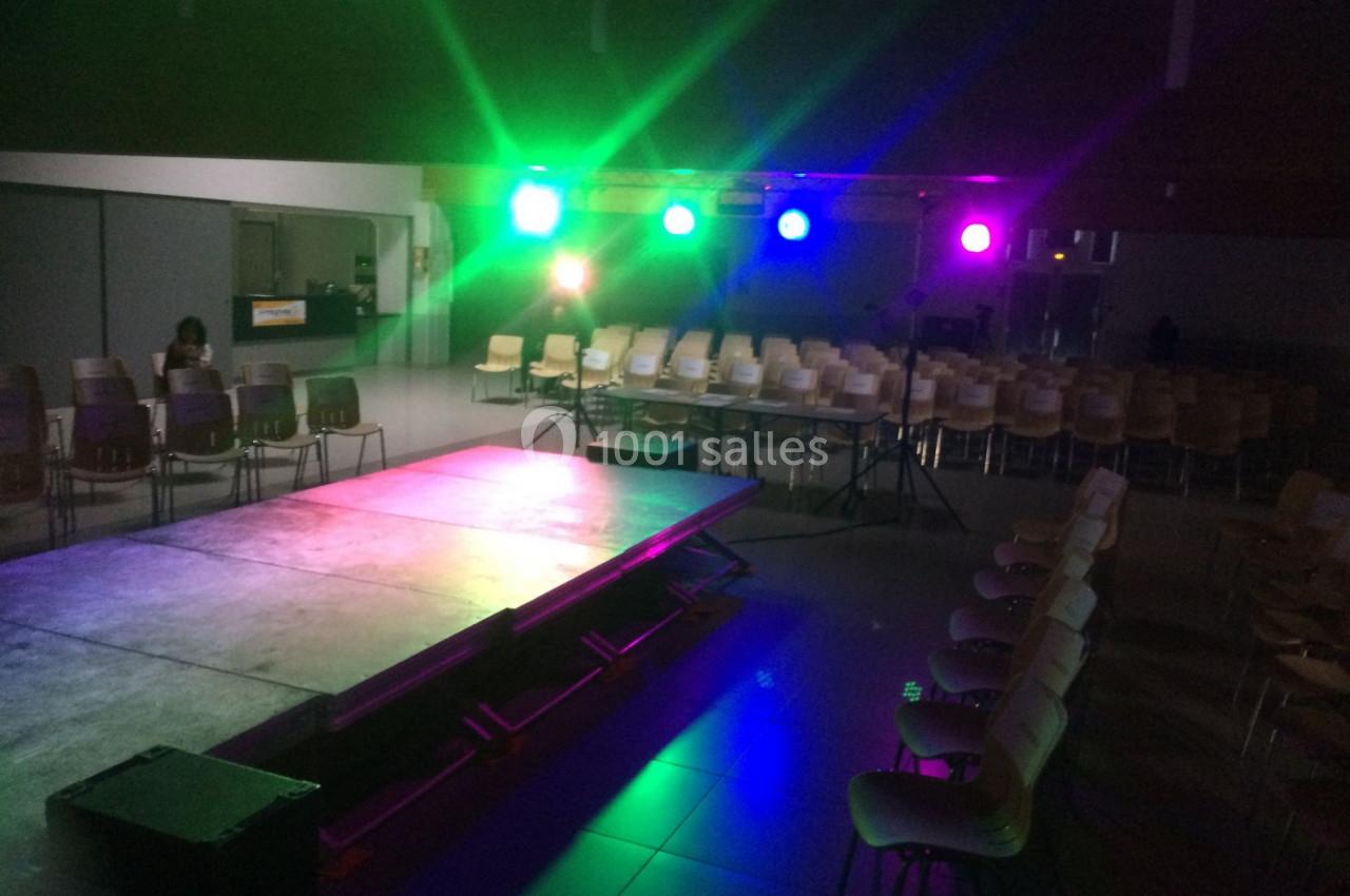 Salle sombre avec une scène éclairée par des projecteurs colorés et des rangées de chaises vides en arrière-plan.
