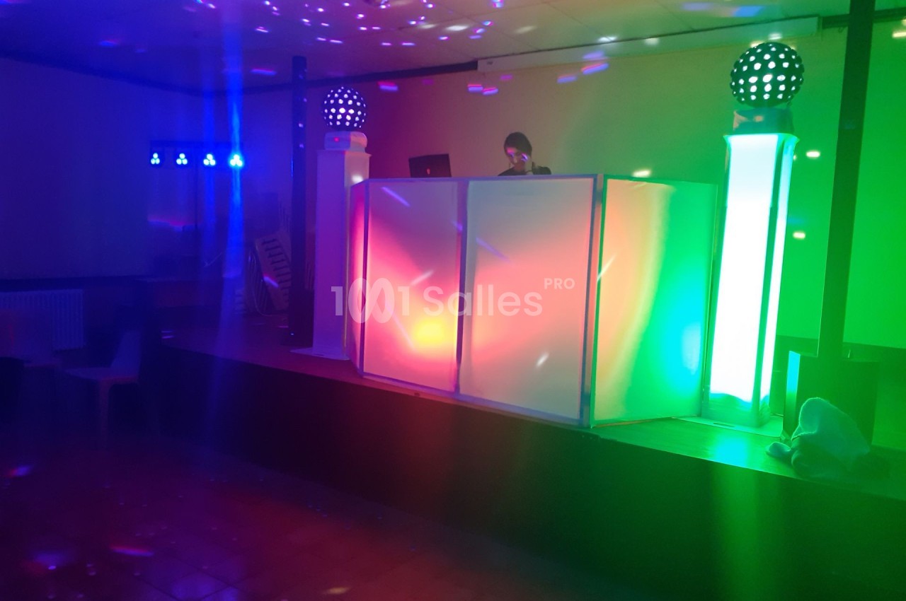 Un DJ mixe derrière une cabine lumineuse aux couleurs vives dans une salle de fête éclairée par des jeux de lumière.