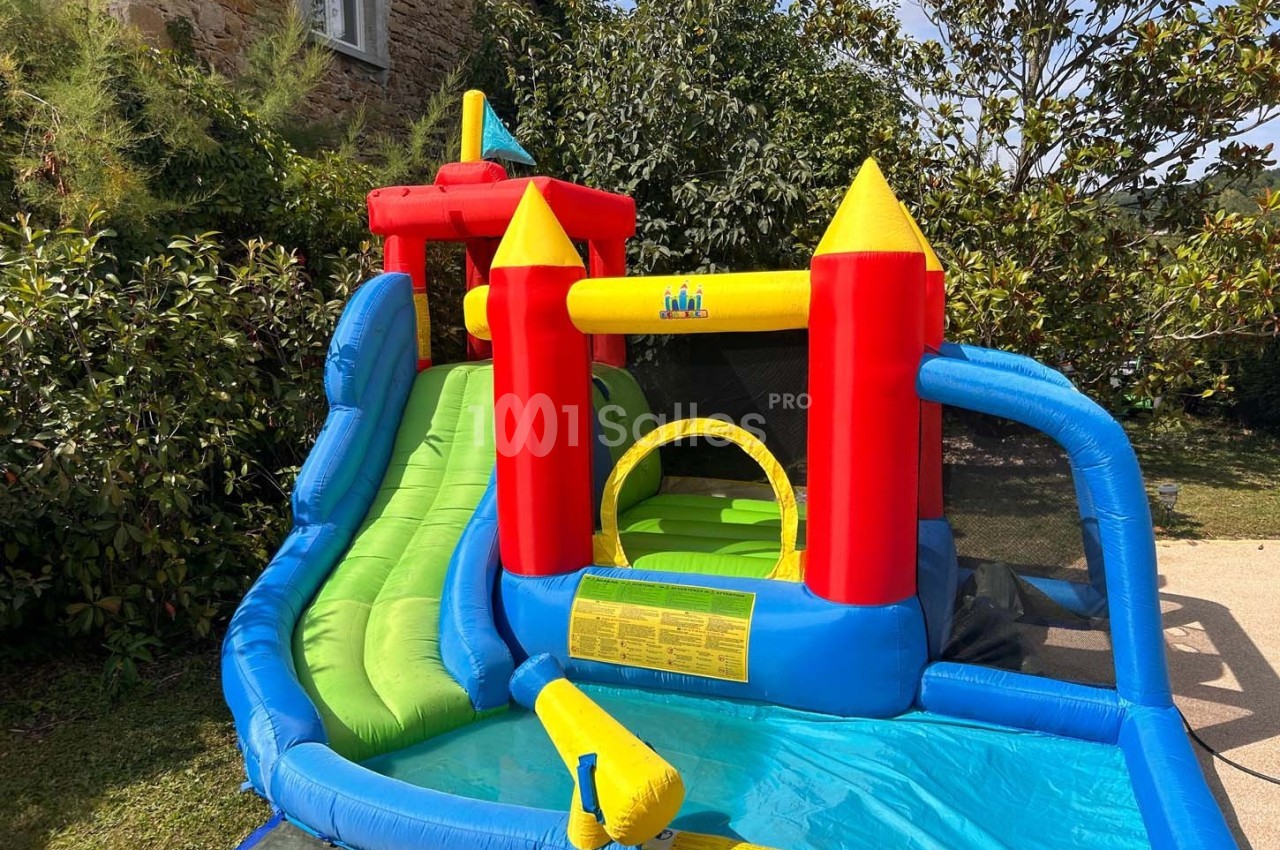 Château gonflable coloré avec toboggan et bassin d'eau, installé dans un jardin verdoyant.