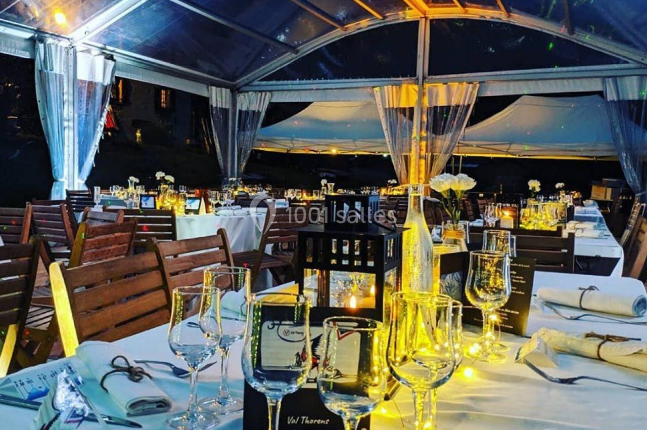 Salle de réception en plein air sous une verrière, avec tables décorées, nappes blanches et éclairage chaleureux.