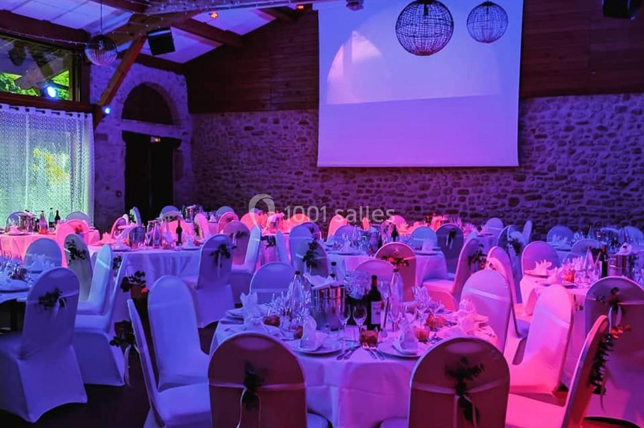Salle de réception décorée avec des tables rondes dressées, éclairage coloré et projecteur en arrière-plan.