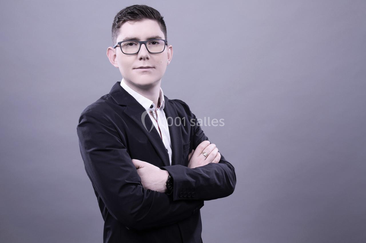Homme en costume sombre, bras croisés, posant devant un fond gris uni.