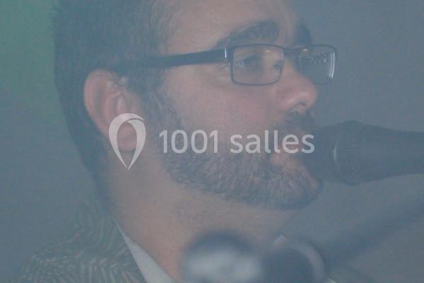 Homme barbu portant des lunettes, chantant dans un microphone dans une ambiance légèrement brumeuse.