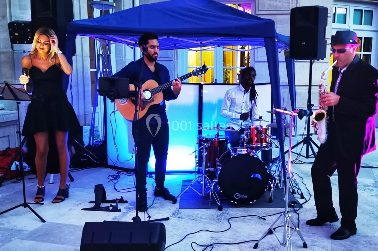 Un groupe de musiciens joue en extérieur sous une tente, avec une chanteuse, un guitariste, un batteur et un saxophoniste.