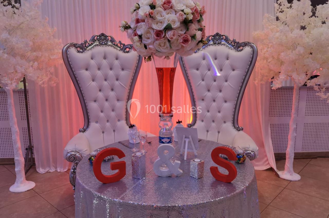 Deux fauteuils blancs capitonnés encadrent une table décorée avec des initiales, des fleurs et un vase rouge.