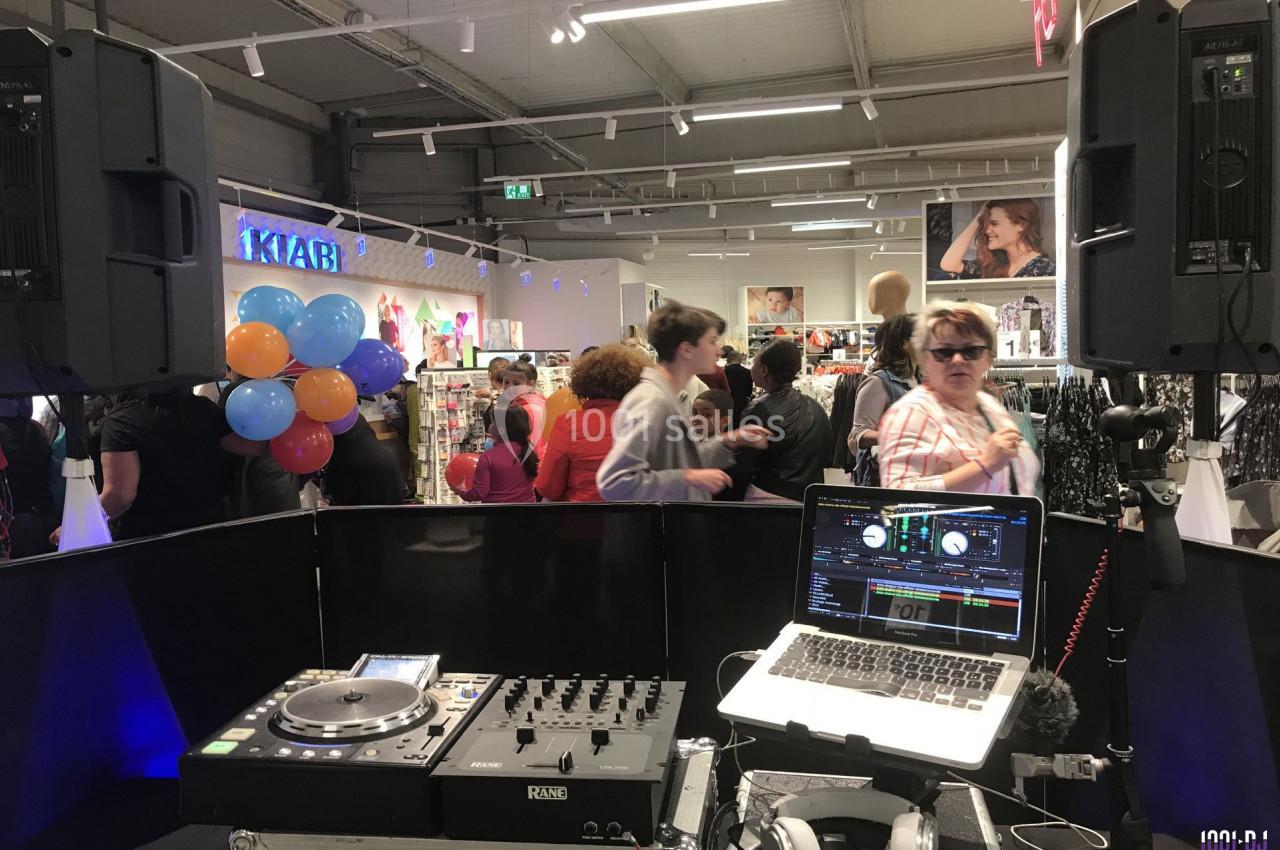 Poste de DJ avec platines et ordinateur dans un magasin, des clients et ballons visibles en arrière-plan.