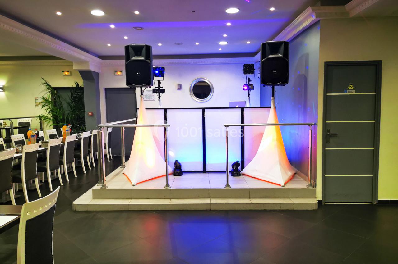 Scène de petite taille équipée de deux enceintes sur pieds, éclairages colorés et tables alignées en arrière-plan.