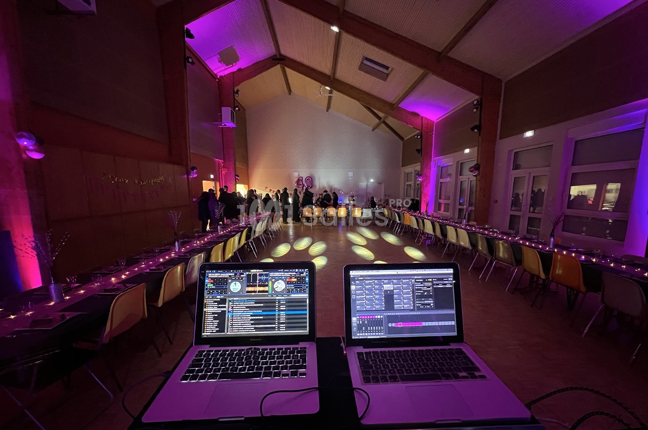 Salle de réception décorée avec des lumières colorées, vue depuis une table de mixage avec deux ordinateurs portables.