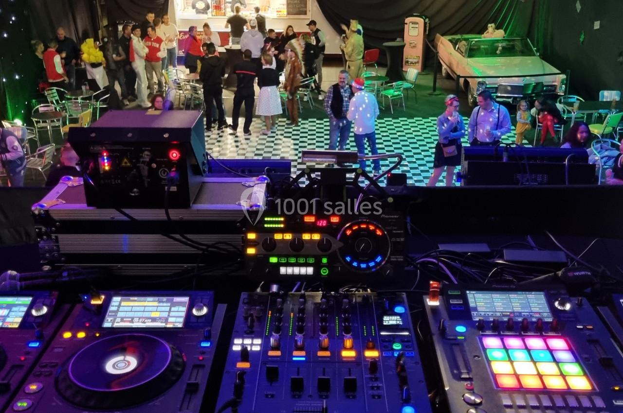 Vue depuis une régie DJ avec platines et table de mixage, face à une salle animée avec des personnes dansant et discutant.