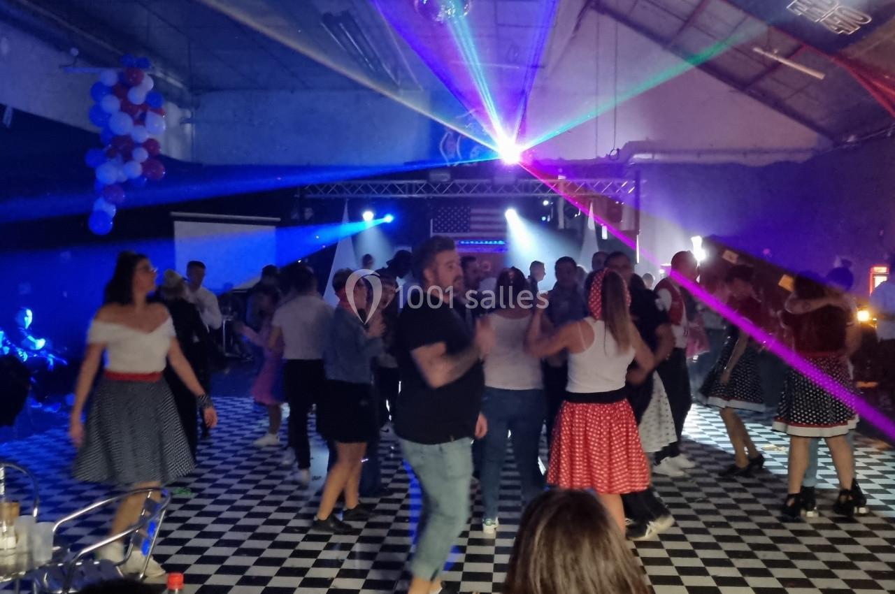 Des personnes dansent sur une piste à damier dans une salle décorée avec des lumières colorées et des tables en arrière-plan.