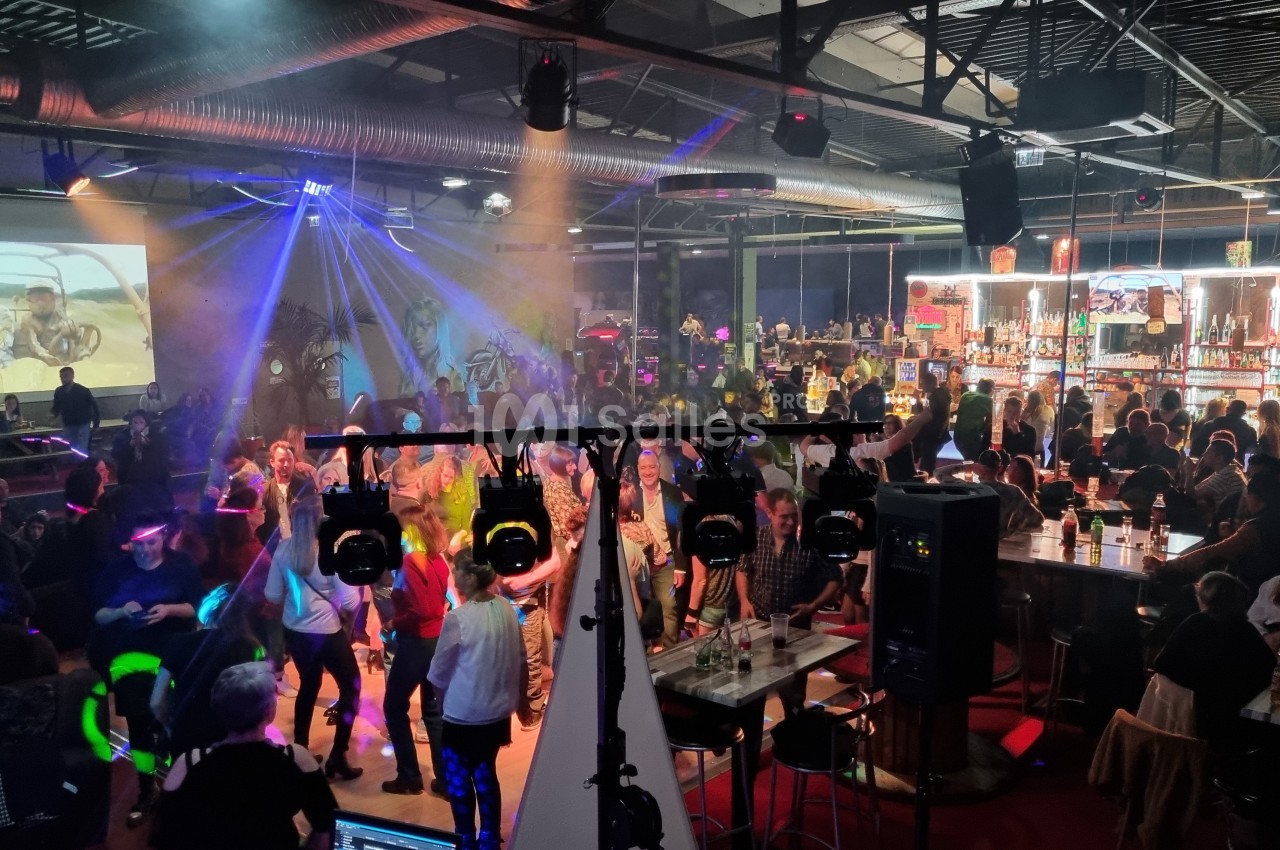 Salle animée avec des personnes dansant et discutant, éclairage coloré, bar illuminé en arrière-plan.