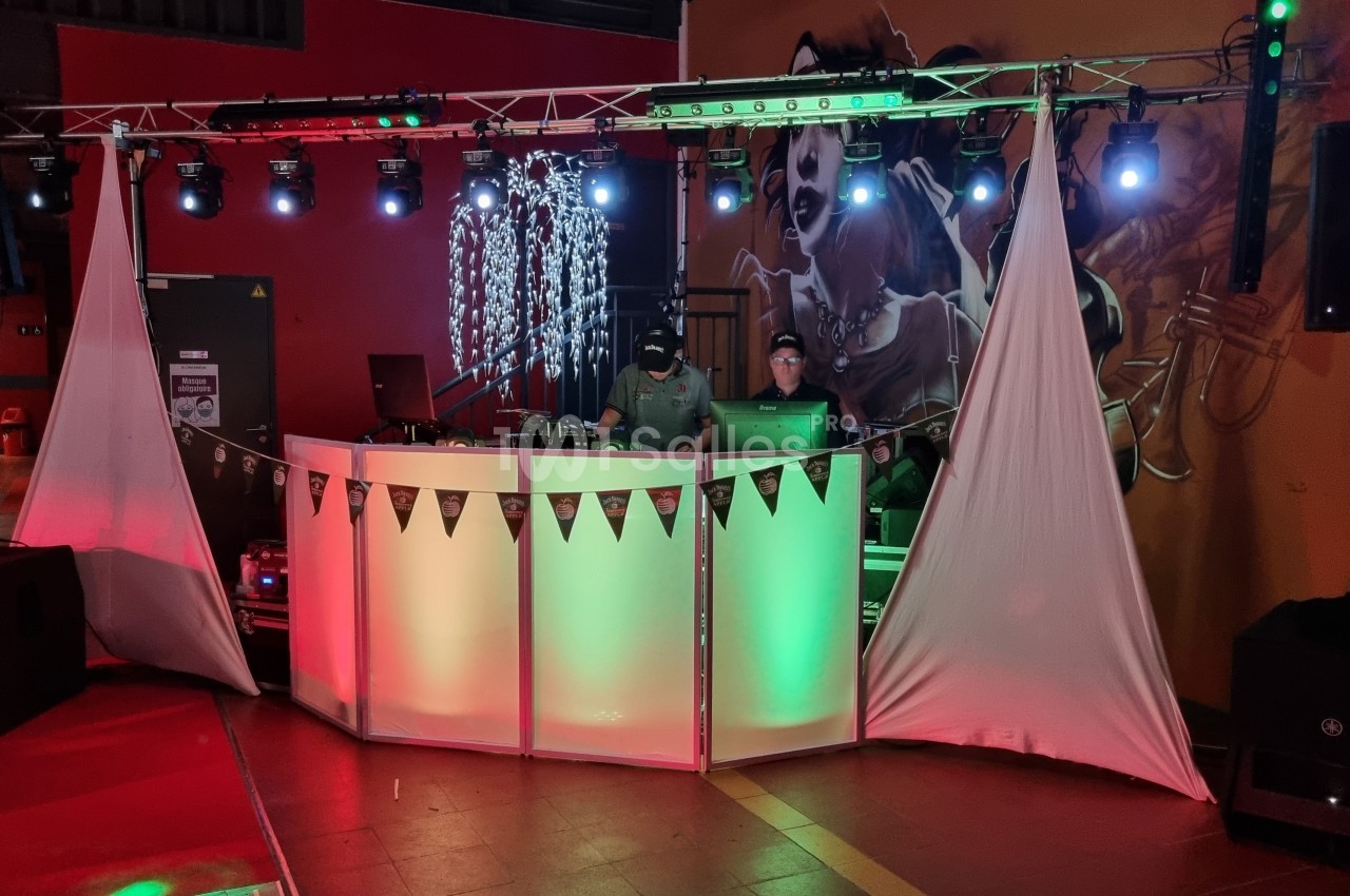Deux DJ mixent derrière une table éclairée, entourée de lumières colorées et de décorations dans une salle sombre.