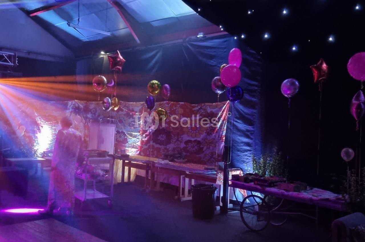 Salle décorée pour une fête avec ballons colorés, lumières tamisées et effets lumineux sur une scène.