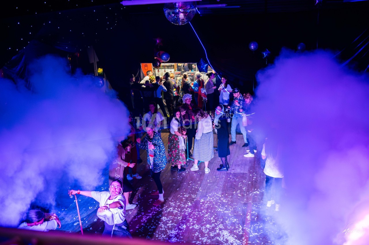 Personnes dansant et socialisant dans une salle festive éclairée par des lumières colorées, avec de la fumée et des…