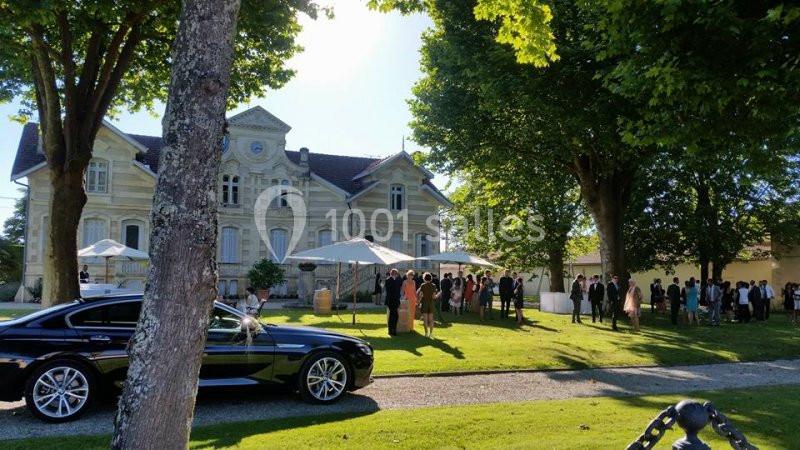 Un groupe de personnes rassemblées devant un manoir entouré d'arbres, avec une voiture noire au premier plan.