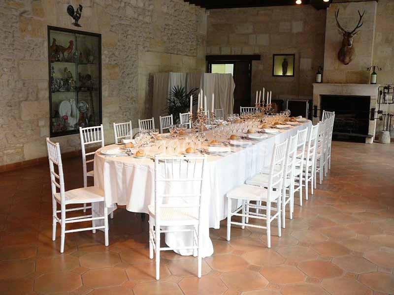 Grande table dressée avec nappes blanches, chaises blanches et chandeliers, dans une salle en pierre avec cheminée.