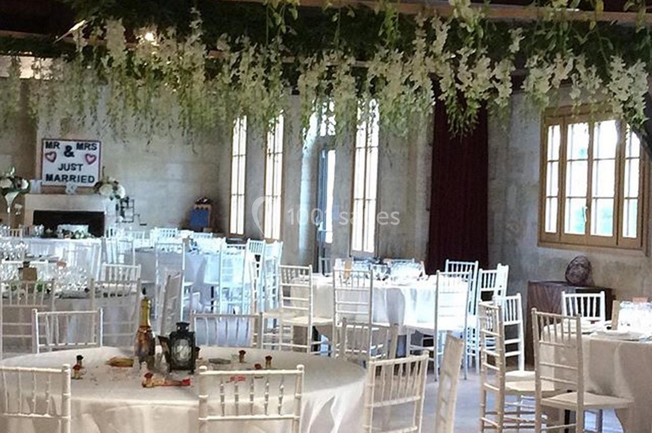 Salle de réception décorée pour un mariage, avec des tables rondes, des chaises blanches et des guirlandes florales…