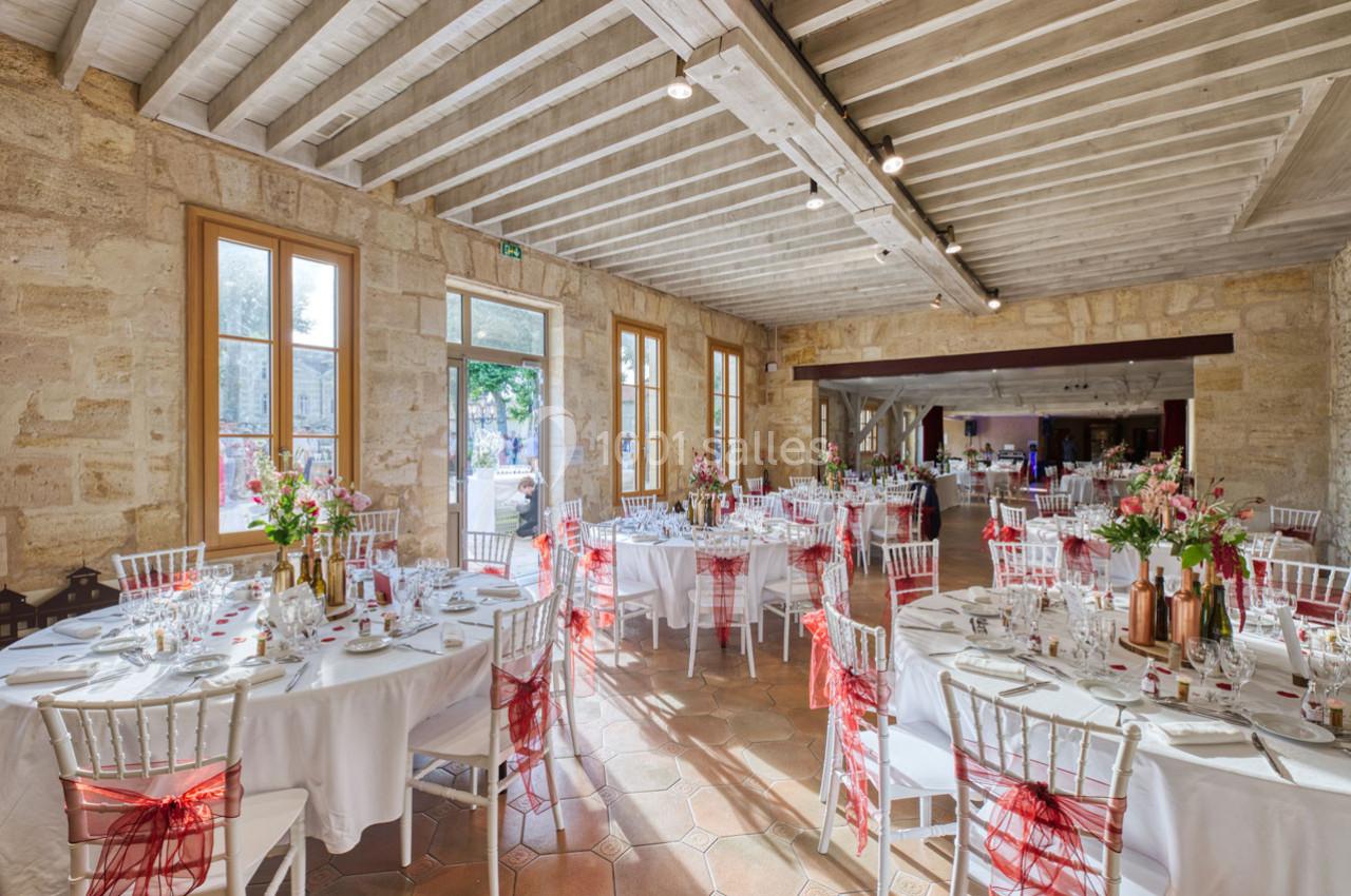 Salle de réception lumineuse avec tables rondes décorées de nappes blanches, chaises ornées de rubans rouges et fleurs.