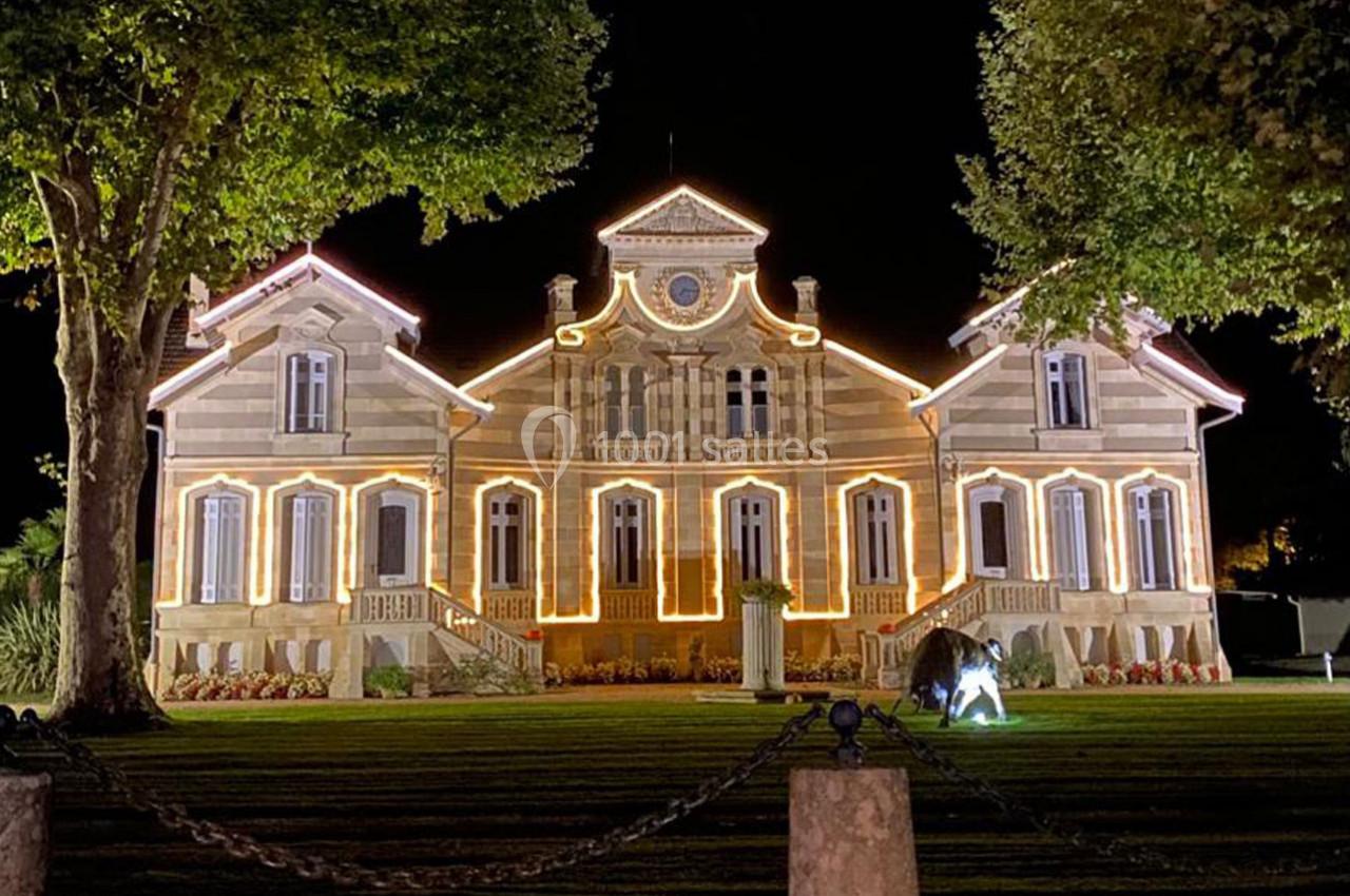 Façade d'un bâtiment éclairé par des guirlandes lumineuses, entouré d'arbres et d'une pelouse, photographié de nuit.